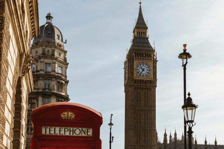 Red telephone booth with Big Ben and historic London architecture in daylight.